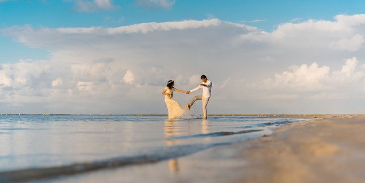 Paar spielt barfuß am Strand und spritzt Wasser im seichten Meer – Ausdruck von Leichtigkeit und Freude während einer Hochzeitsreise.