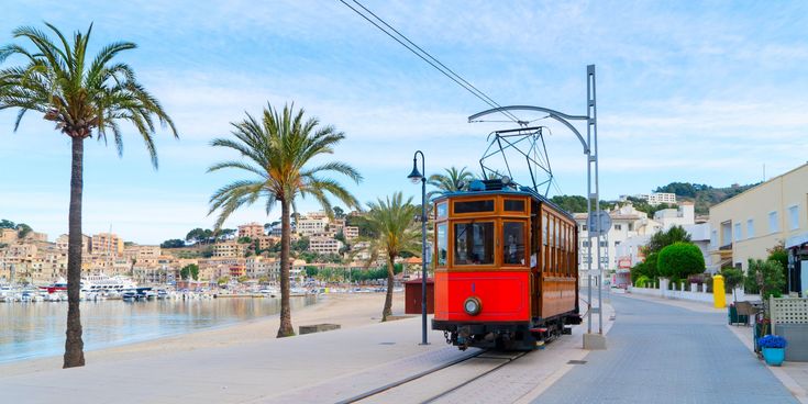 Historische Straßenbahn ‚Roter Blitz‘ fährt an der Strandpromenade von Port de Sóller auf Mallorca entlang, gesäumt von Palmen und Hafenblick.
