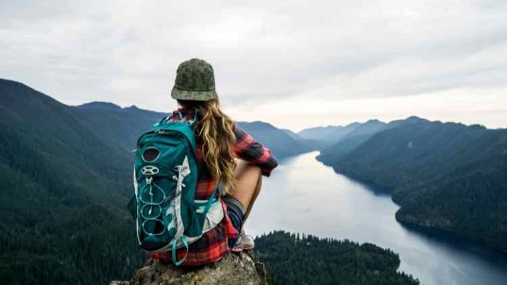 Frau mit Rucksack sitzt auf Stein mit Aussicht auf ein Tal mit Wasser 