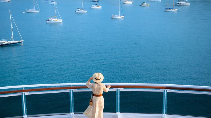 Frau in elegantem Sommerkleid und Strohhut steht an der Reling eines Kreuzfahrtschiffs und blickt auf eine Bucht mit Segelbooten – Moment der Ruhe und Freiheit auf See.