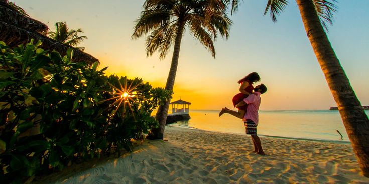 Paar umarmt sich am Strand unter Palmen bei Sonnenuntergang – romantischer Moment im goldenen Abendlicht am Meer.