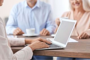 Person arbeitet an einem Laptop, während zwei weitere Menschen im Hintergrund bei einer Tasse Kaffee sitzen und sich unterhalten.