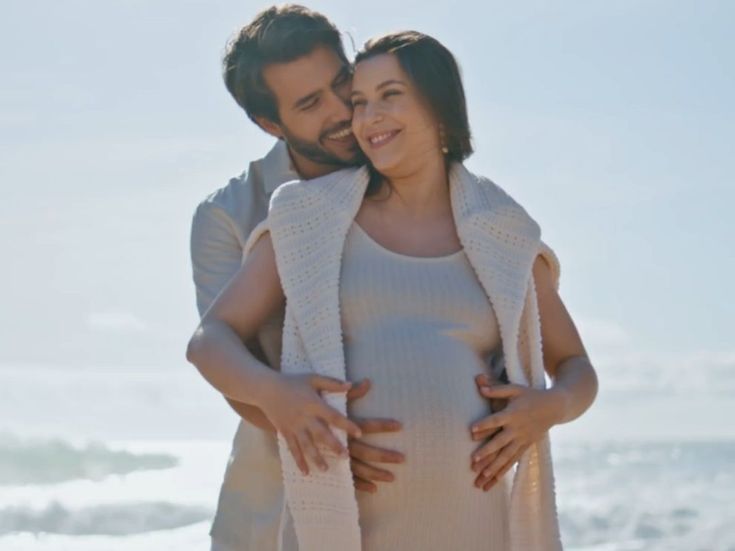 Glückliches Paar am Meer, der Mann umarmt die schwangere Frau liebevoll – harmonische Babymoon-Stimmung in sanftem Licht.