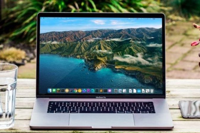 Laptop auf einem Holztisch im Garten mit Glas Wasser, Notizblock und Smartphone – Symbol für mobiles Arbeiten im Freien.