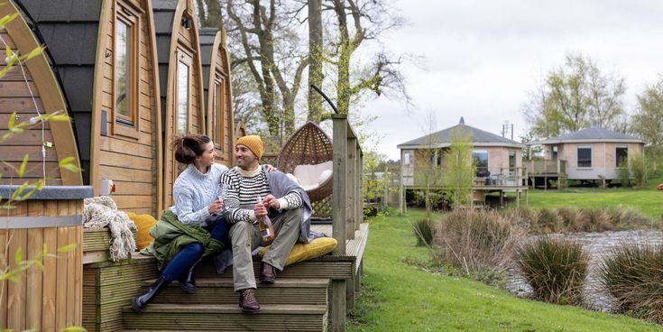 Paar sitzt mit Getränken auf der Terrasse eines Glamping-Häuschens aus Holz, umgeben von Natur und kleinen Teichen.