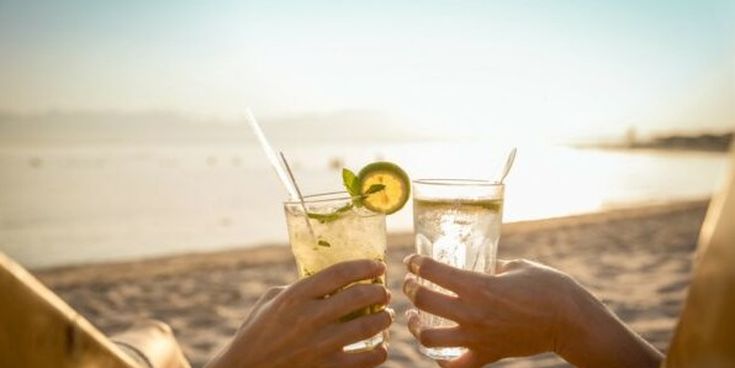Zwei Personen stoßen mit kühlen Getränken am Strand bei Sonnenuntergang an – Symbol für Entspannung und Urlaubsfeeling.