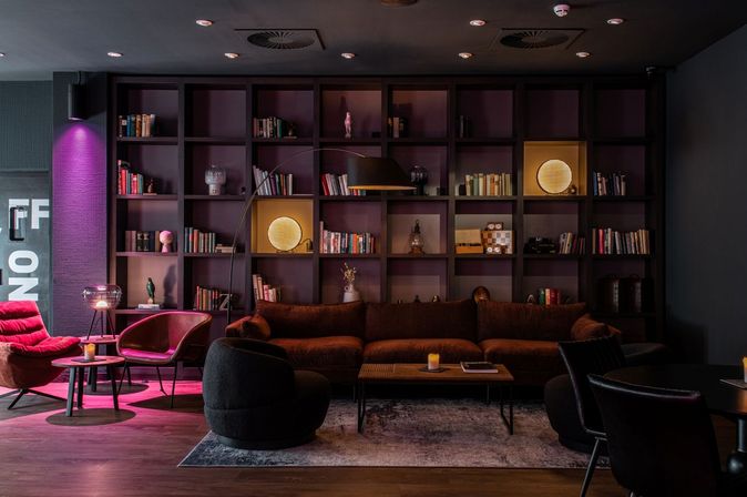 Gemütliche Lounge des Penta Hotels mit Sesseln, Sofa, Wandregal im Hintergrund und stimmungsvoller Beleuchtung 