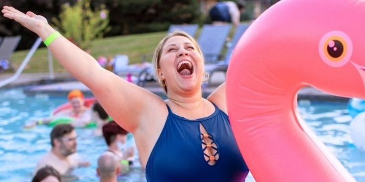 Lachende Frau im blauen Badeanzug sitzt mit ausgebreiteten Armen auf einem pinken Flamingo im Pool.