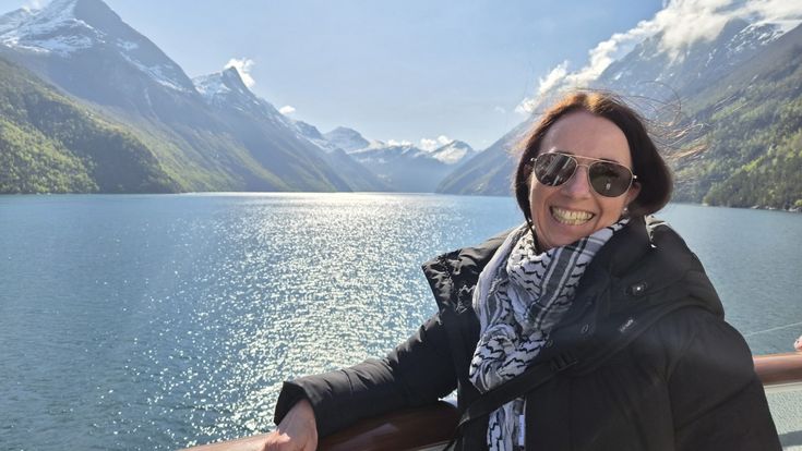 Sandra Janissen lächelt an der Reling eines Schiffes vor einem glitzernden Fjord und schneebedeckten Bergen.