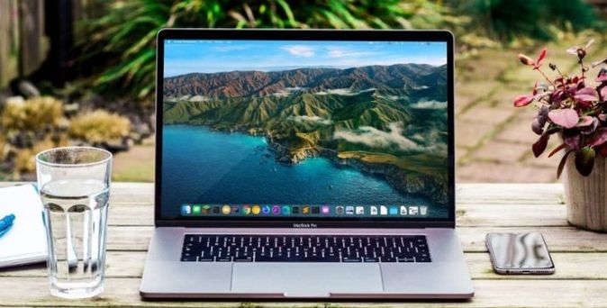 Laptop auf einem Holztisch im Garten mit Glas Wasser, Notizblock und Smartphone – Symbol für mobiles Arbeiten im Freien.