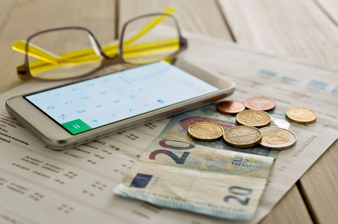 Brille, Smartphone mit Taschenrechner-Display, Euro-Münzen und 20-Euro-Schein liegen auf einem Tisch