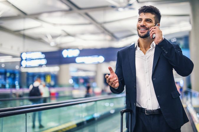Lächelnder Geschäftsmann mit Rollkoffer telefoniert in einem modernen Flughafenterminal.