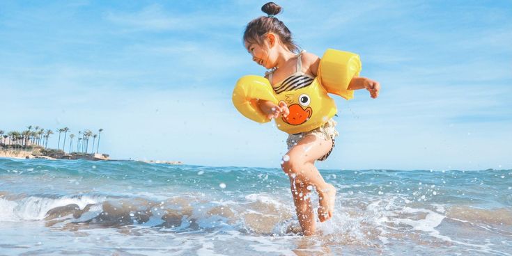 Mädchen mit gelben Schwimmhilfen läuft lachend durch die Wellen am sonnigen Strand.