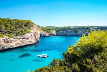 Eine kleine Bucht auf Mallorca mit türkisblauem Wasser und einem Boot, umgeben von felsigen Klippen und grüner Vegetation unter klarem Himmel.