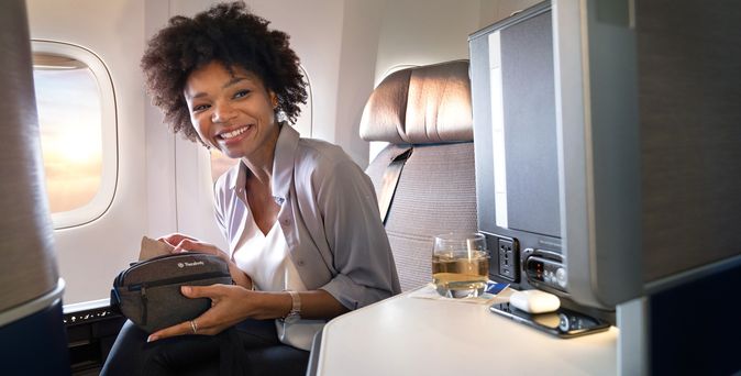 Lächelnde Frau sitzt komfortabel in der Business Class eines Flugzeugs mit einem Getränk und einer Reisetasche.