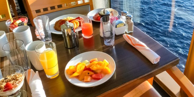 Frühstückstisch mit frischem Obst, Müsli und Säften am Fenster eines Kreuzfahrtschiffs mit Blick auf das blaue Meer.