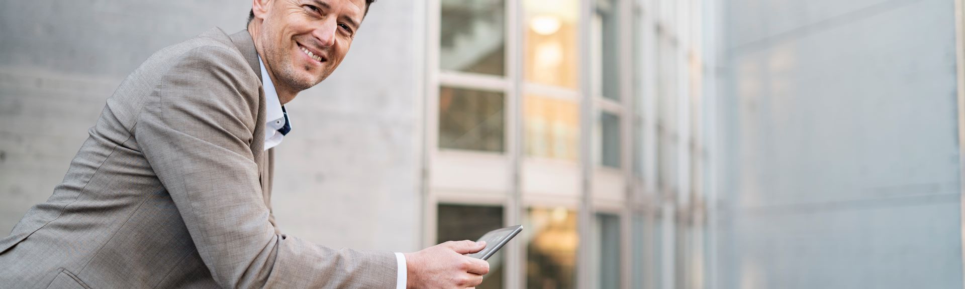 Geschäftsmann lächelt freundlich und lehnt an einem Geländer und hat ein Tablet in der Hand.