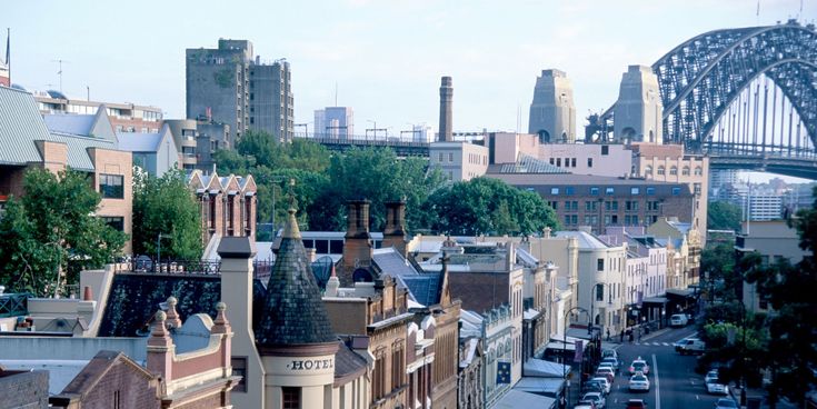 Straßenszene im historischen Viertel The Rocks in Sydney mit alten Backsteingebäuden, Hotels und Blick auf die Sydney Harbour Bridge im Hintergrund.