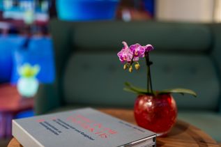 Nahaufnahme einer violetten Orchidee in roter Vase neben einem Buch mit dem Titel ‚Reise in die Antarktis‘ auf einem Holztisch.