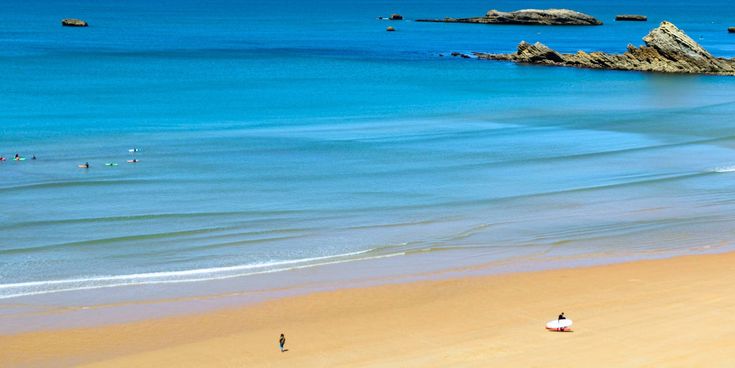 Weiter Sandstrand an der Côte d’Argent in Frankreich mit ruhigem türkisfarbenem Atlantik, sanften Wellen und einzelnen Surfern im Wasser; im Hintergrund ragen Felsen aus dem Meer.