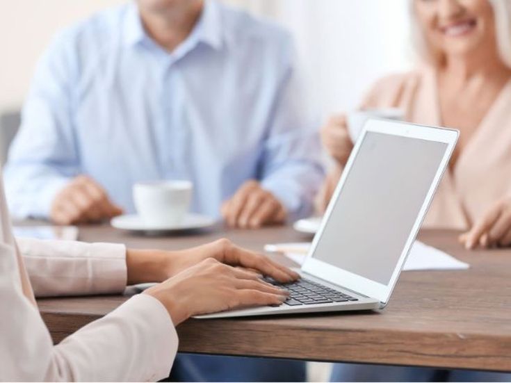 Person arbeitet an einem Laptop, während zwei weitere Menschen im Hintergrund bei einer Tasse Kaffee sitzen und sich unterhalten.