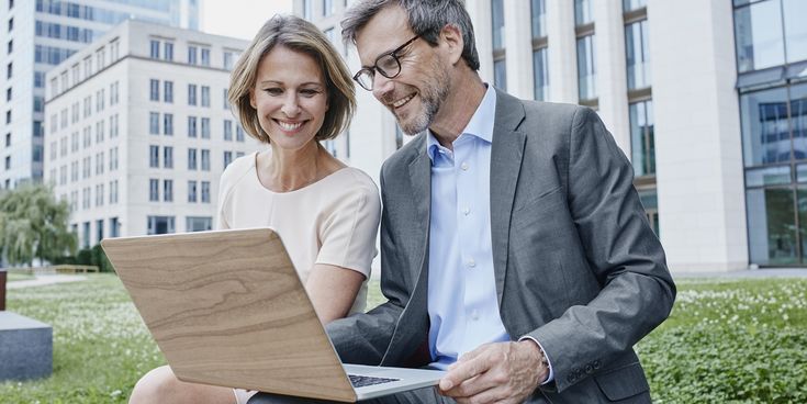 Ein lächelnder Mann und eine Frau in Business-Kleidung sitzen im Grünen vor modernen Bürogebäuden und schauen gemeinsam auf einen Laptop