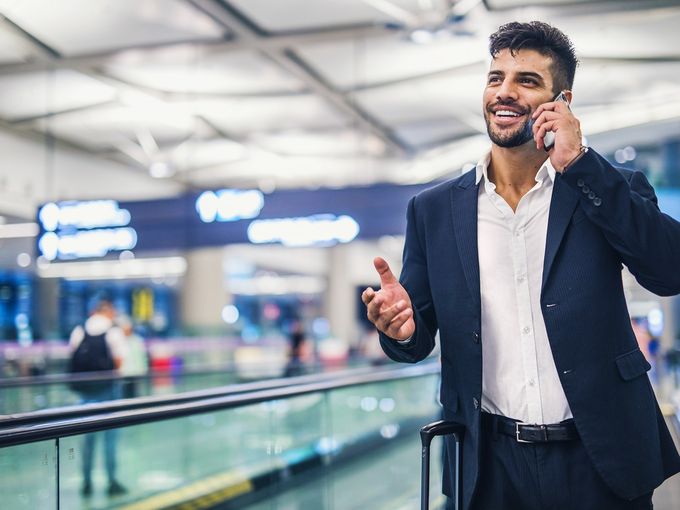 Lächelnder Geschäftsmann mit Rollkoffer telefoniert in einem modernen Flughafenterminal.