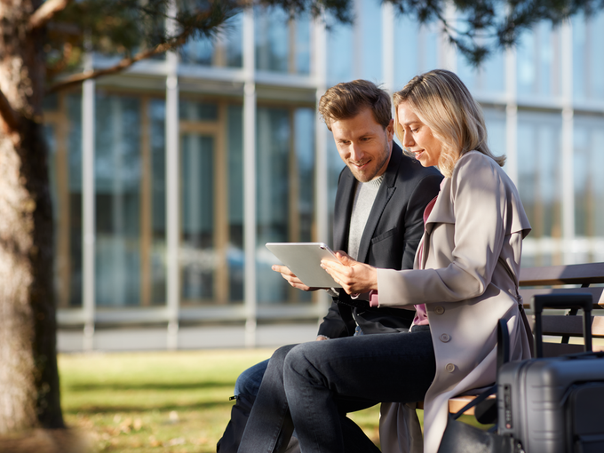 Geschäftsreisende (Mann und Frau) auf einer Bank vor einem Bürogebäude mit einem Tablet in der Hand