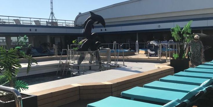 Auf dem Sonnendeck eines Kreuzfahrtschiffs stehen mehrere türkisfarbene Liegen in der Sonne. In der Mitte befindet sich ein Poolbereich mit einer Skulptur von zwei springenden Delfinen. Einige Pflanzen und Passagiere sind im Hintergrund zu sehen, während der Himmel klar und blau ist.
