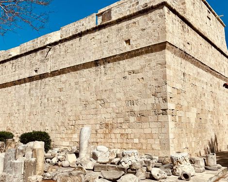 Massive Steinmauer eines historischen Gebäudes mit Ruinen aus zerbrochenen Säulen und Steinen im Vordergrund unter blauem Himmel.