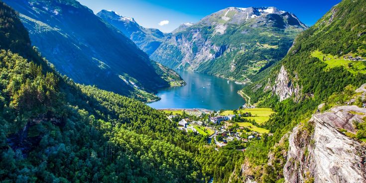 Luftaufnahme des Geirangerfjords in Norwegen mit türkisblauem Wasser, umgeben von steilen, bewaldeten Bergen und schneebedeckten Gipfeln unter klarem, blauem Himmel.