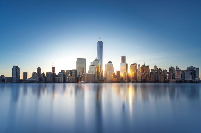 Skyline von New York mit One World Trade Center bei Sonnenuntergang, Spiegelung im Wasser.