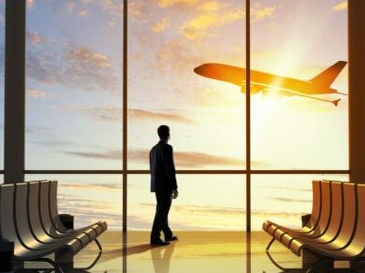 Reisender beobachtet durch die Glasfassade eines Flughafens den Start eines Flugzeugs bei Sonnenuntergang.