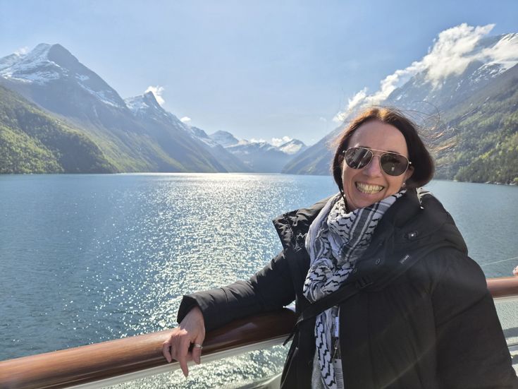 Sandra Janissen lächelt an der Reling eines Schiffes vor einem glitzernden Fjord und schneebedeckten Bergen.