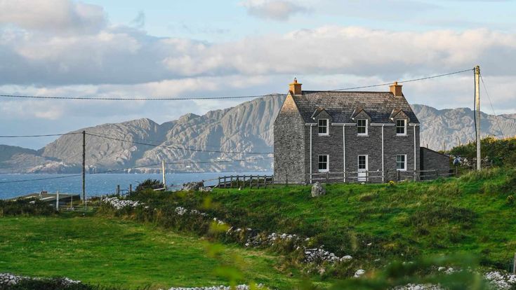 Einsames Steinhaus inmitten grüner Wiesen mit Bergen im Hintergrund.