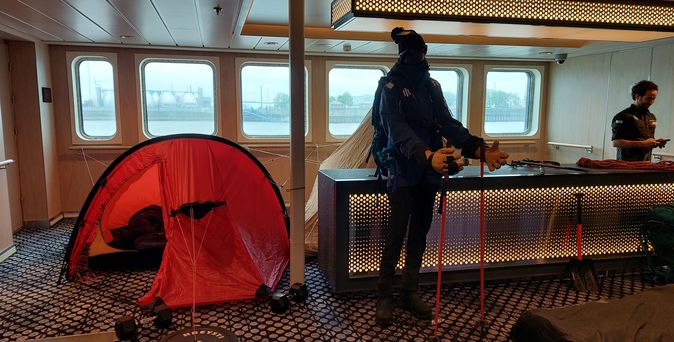 Präsentation von Polarausrüstung an Bord: Eine Person trägt warme Expeditionskleidung und steht neben einem roten Zelt und Schlafsäcken.