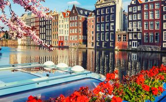Bunte Giebelhäuser am Kanal in Amsterdam mit Frühlingsblüten und Blumen am Boot unter blauem Himmel.