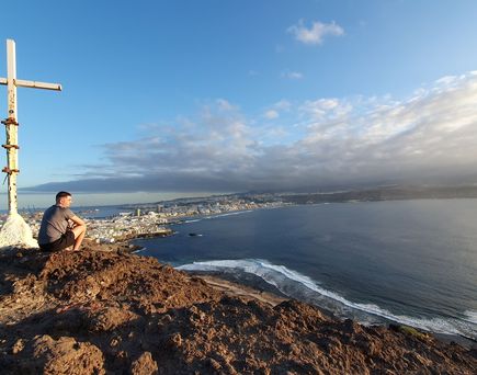 Ein junger Mann sitzt in der Abendsonne auf einem Felsen vor einem großen weißen Kreuz und blickt auf den Strand von Las Palmas auf Gran Canaria