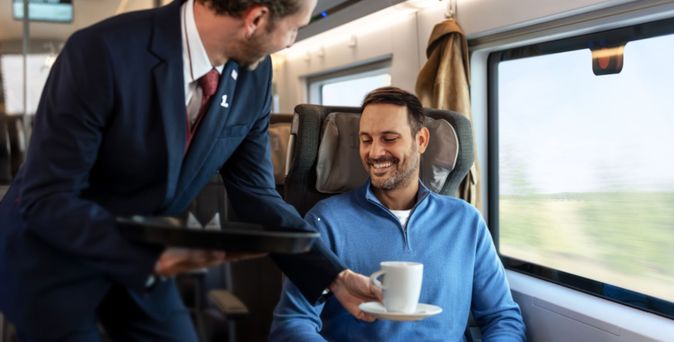 Zugbegleiter serviert einem lächelnden Fahrgast im 1.-Klasse-Abteil Kaffee, am Tisch liegt ein Tablet.