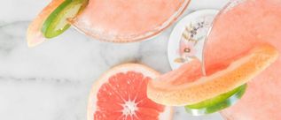 Zwei rosafarbene Grapefruit-Cocktails mit Zitrusscheiben und Gurke auf einer hellen Marmorfläche.