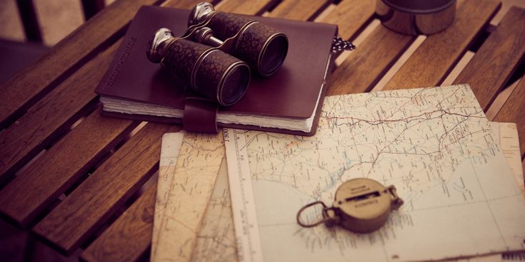 Vintage-Fernglas, Leder-Notizbuch, alte Landkarten und Kompass liegen auf einem Holztisch – Symbol für Abenteuerlust und Reiseplanung.