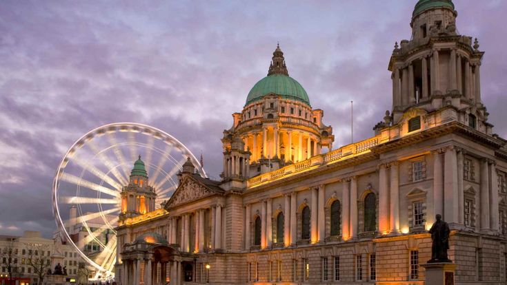 Beleuchtetes Regierungsgebäude mit Riesenrad im Hintergrund bei Abendstimmung in Belfast.