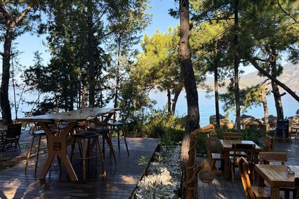 Holzterrassen mit Tischen und Stühlen unter Kiefern, im Hintergrund Meer und blauer Himmel.