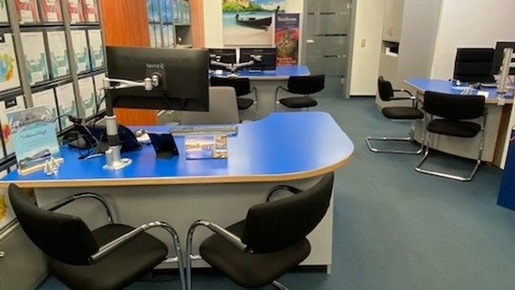 Innenansicht eines Reisebüros mit mehreren Beratungsplätzen, blauen Schreibtischen und Prospektwand.