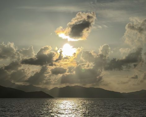 Tief stehende Sonne zwischen dramatischen Wolken über dem Meer, mit dunkler Inselsilhouette am Horizont.