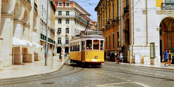 Gelbe Straßenbahn fährt durch eine Straße mit historischen Gebäuden und Oberleitungen in der Innenstadt von Lissabon.