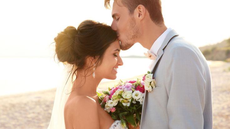 Ein frisch verheiratetes Paar steht am Strand im warmen Sonnenlicht. Die Braut hält einen bunten Blumenstrauß und lächelt, während der Bräutigam ihr sanft einen Kuss auf die Stirn gibt. Beide tragen festliche Hochzeitskleidung.