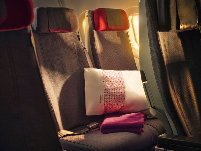 TAP-Economy-Prime-Sitz mit weißem Kissen mit rotem Muster und pinkfarbener Decke, im warmen Licht des Flugzeugfensters.