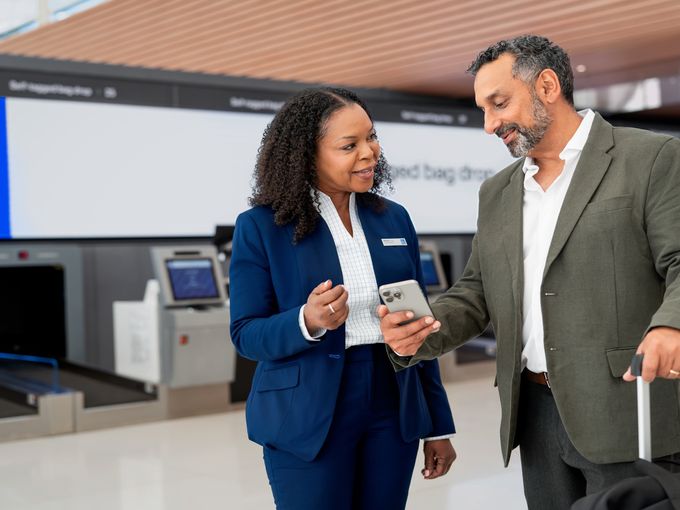 Eine Mitarbeiterin im blauen Blazer zeigt einem Reisenden mit Koffer etwas auf seinem Smartphone, im Hintergrund Self-Service-Check-in-Automaten.