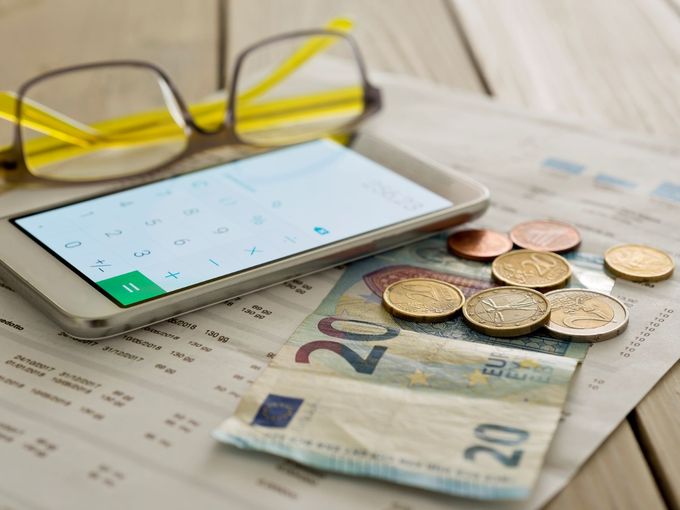 Brille, Smartphone mit Taschenrechner-Display, Euro-Münzen und 20-Euro-Schein liegen auf einem Tisch